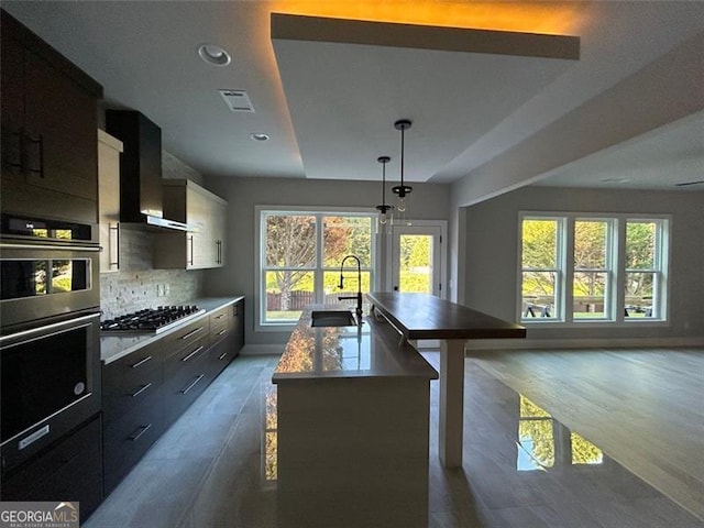 kitchen featuring sink, decorative light fixtures, a center island with sink, appliances with stainless steel finishes, and wall chimney range hood