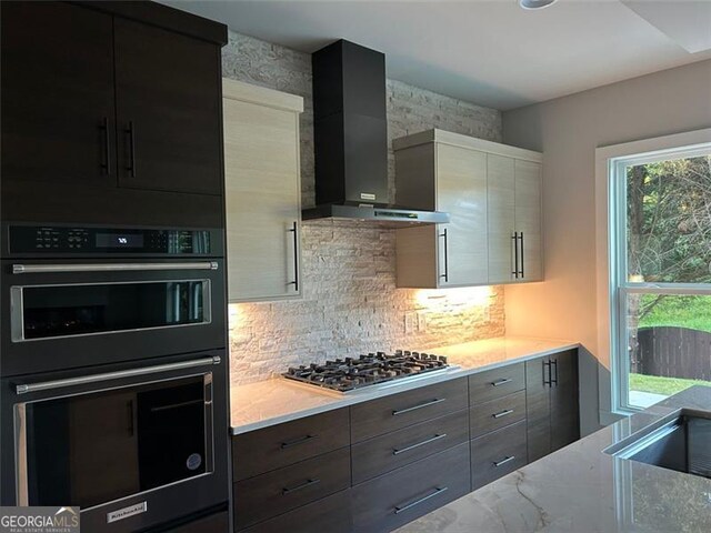 kitchen with double oven, stainless steel gas stovetop, light stone countertops, decorative backsplash, and wall chimney range hood