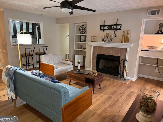 living room with ceiling fan, a fireplace, a textured ceiling, built in shelves, and light wood-type flooring