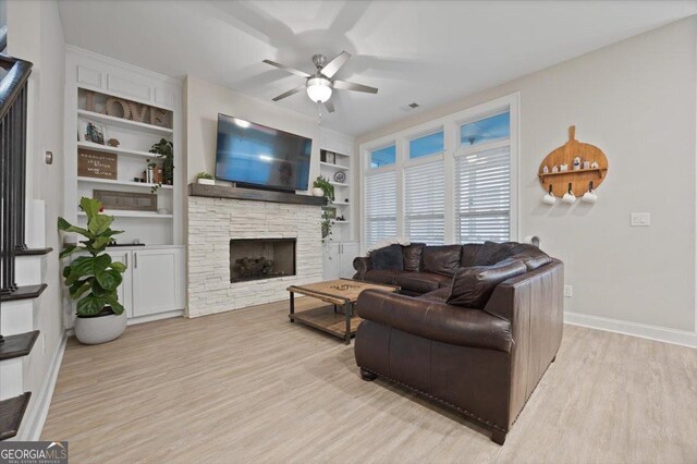 living room with ceiling fan, a stone fireplace, light hardwood / wood-style floors, and built in shelves