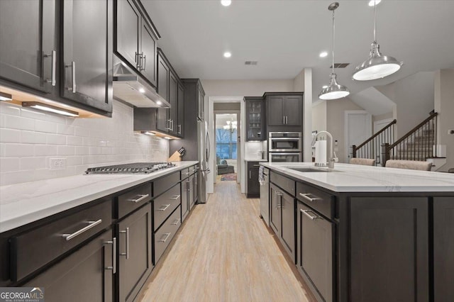 kitchen with sink, light hardwood / wood-style flooring, appliances with stainless steel finishes, a kitchen island with sink, and hanging light fixtures