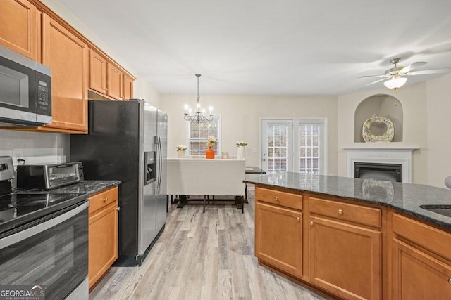 kitchen with ceiling fan with notable chandelier, pendant lighting, dark stone countertops, stainless steel appliances, and light hardwood / wood-style flooring