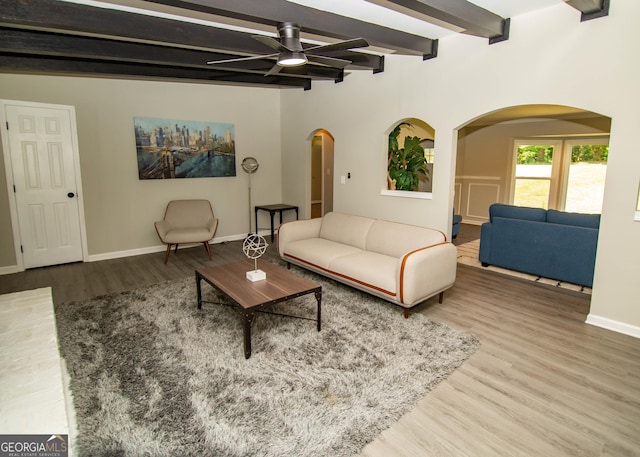 living room featuring baseboards, beamed ceiling, wood finished floors, arched walkways, and a ceiling fan