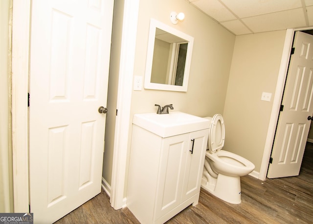 half bathroom with vanity, wood finished floors, baseboards, a paneled ceiling, and toilet