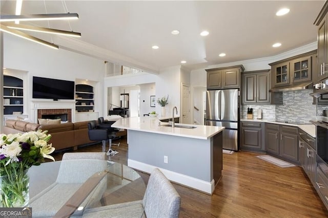 kitchen with sink, dark wood-type flooring, stainless steel refrigerator, an island with sink, and a brick fireplace