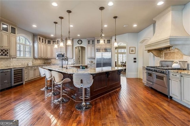 kitchen with wine cooler, custom exhaust hood, decorative light fixtures, a center island with sink, and premium appliances