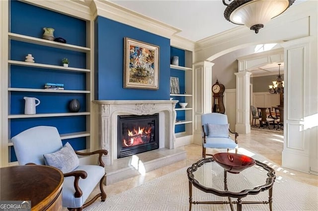 living area with built in features, a premium fireplace, an inviting chandelier, decorative columns, and ornamental molding