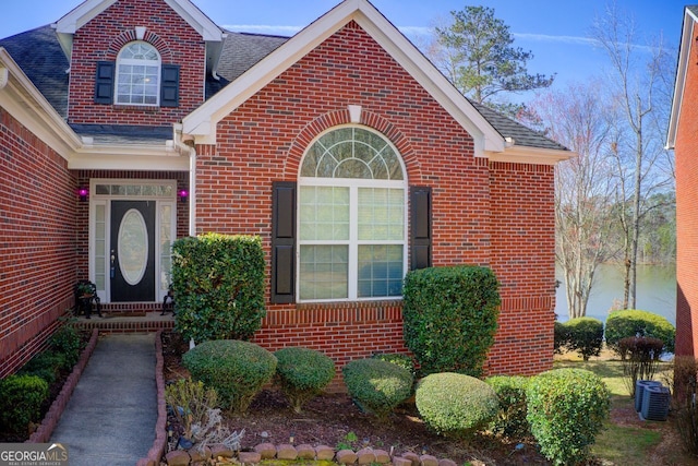 property entrance featuring brick siding, cooling unit, and a shingled roof