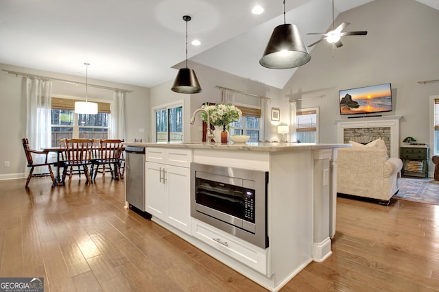 kitchen featuring a stone fireplace, stainless steel microwave, white cabinets, hanging light fixtures, and a kitchen island with sink