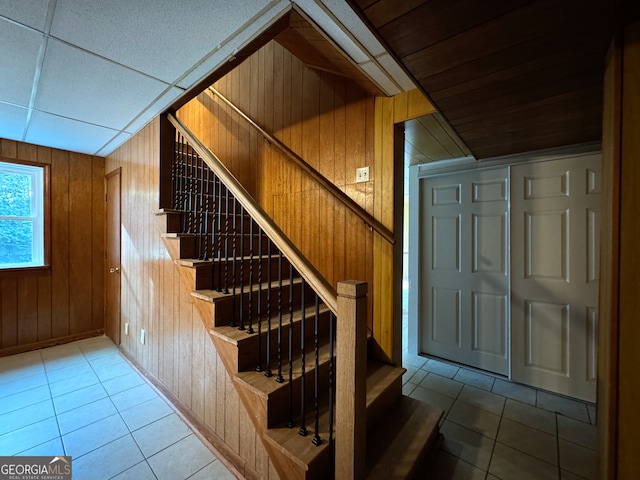 staircase featuring tile patterned flooring, a drop ceiling, and wood walls