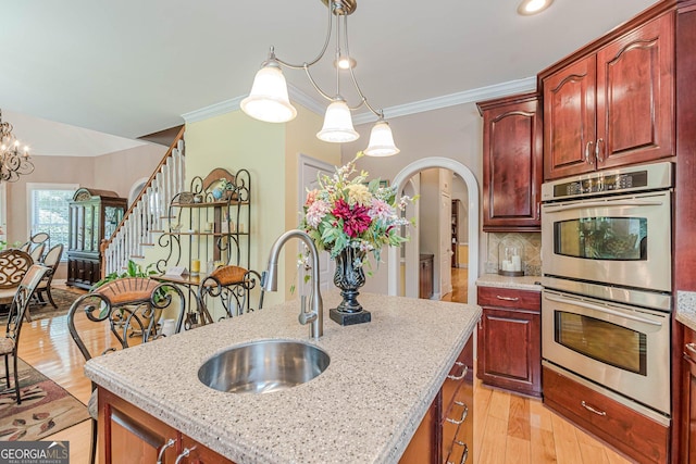 kitchen featuring pendant lighting, an island with sink, sink, light stone counters, and stainless steel double oven