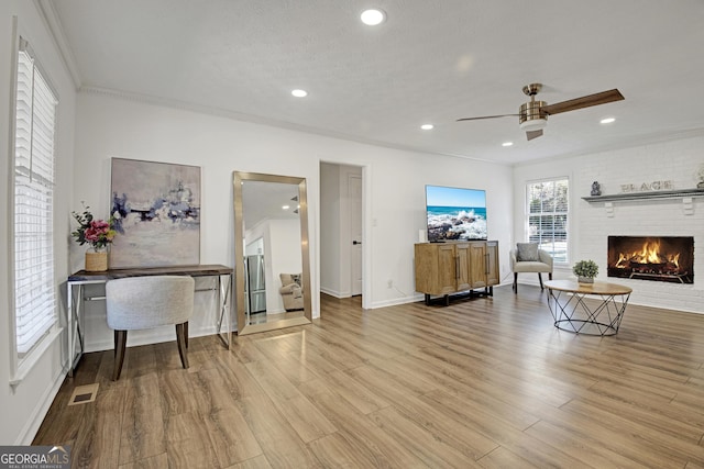 living room featuring ornamental molding, ceiling fan, light hardwood / wood-style floors, a brick fireplace, and a textured ceiling