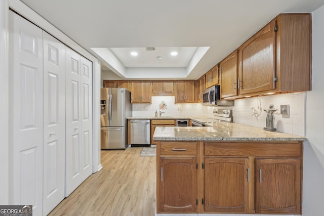 kitchen with appliances with stainless steel finishes, a tray ceiling, tasteful backsplash, kitchen peninsula, and light wood-type flooring