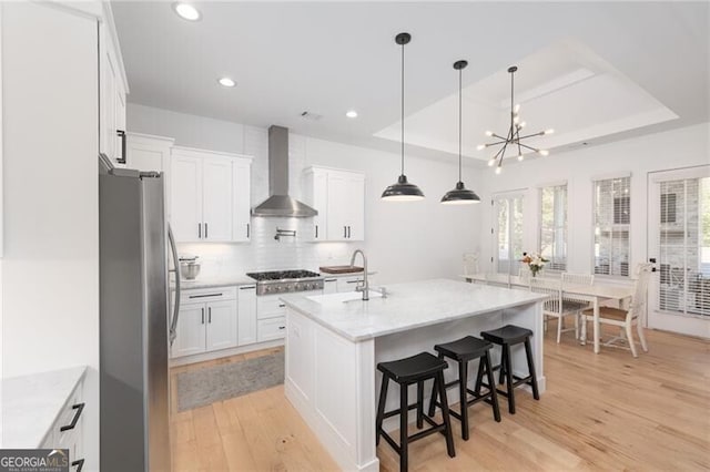 kitchen featuring wall chimney range hood, sink, appliances with stainless steel finishes, a tray ceiling, and white cabinets