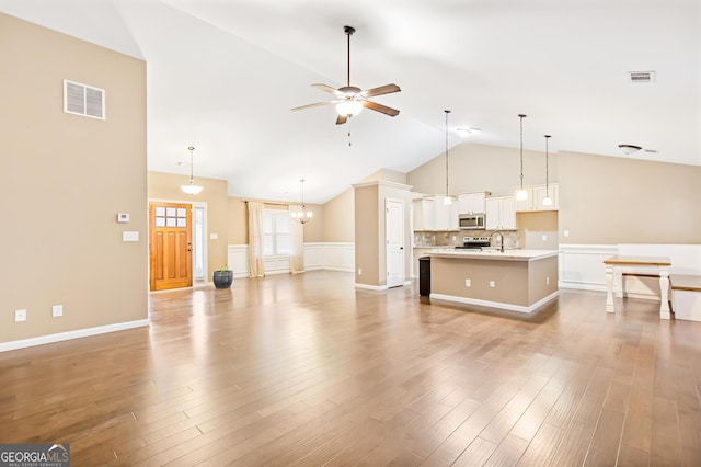 unfurnished living room with sink, ceiling fan with notable chandelier, high vaulted ceiling, and light wood-type flooring