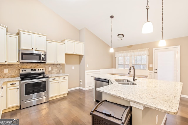 kitchen with an island with sink, sink, hanging light fixtures, stainless steel appliances, and light stone countertops