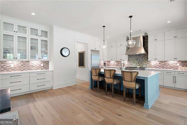 kitchen featuring pendant lighting, wall chimney range hood, a breakfast bar, white cabinetry, and a center island with sink