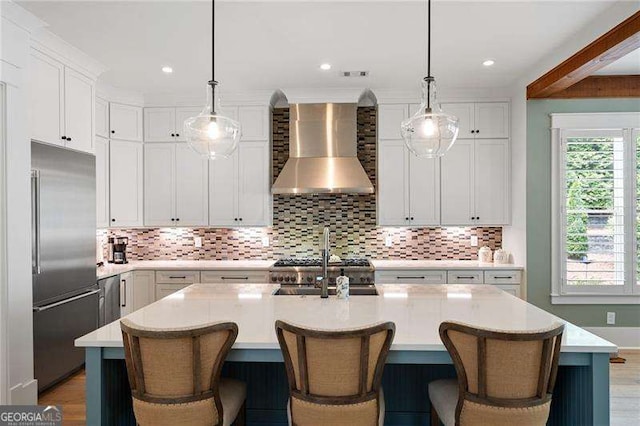 kitchen with appliances with stainless steel finishes, a kitchen island with sink, wall chimney range hood, and decorative light fixtures