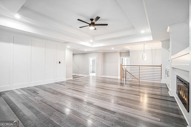 unfurnished living room featuring a decorative wall, wood finished floors, ornamental molding, a raised ceiling, and a glass covered fireplace