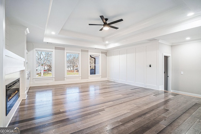 unfurnished living room featuring a raised ceiling, ornamental molding, a glass covered fireplace, ceiling fan, and hardwood / wood-style floors