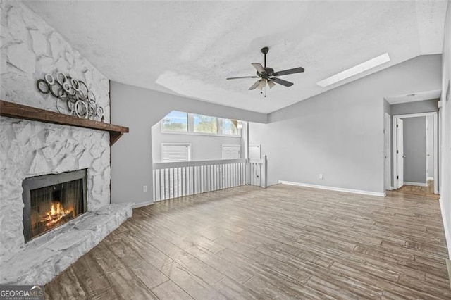 unfurnished living room with ceiling fan, lofted ceiling with skylight, wood-type flooring, a textured ceiling, and a stone fireplace