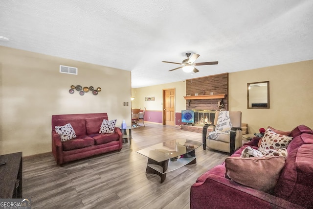 living room featuring a brick fireplace, hardwood / wood-style flooring, a textured ceiling, and ceiling fan