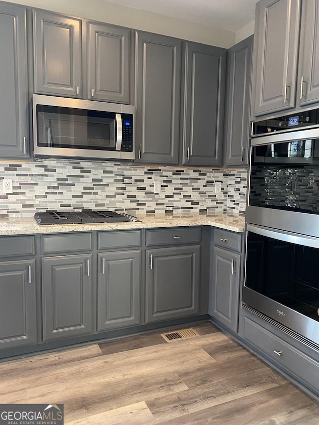 kitchen with appliances with stainless steel finishes, gray cabinetry, decorative backsplash, light stone counters, and light hardwood / wood-style flooring