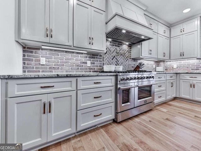 kitchen with custom exhaust hood, tasteful backsplash, light hardwood / wood-style flooring, light stone countertops, and range with two ovens