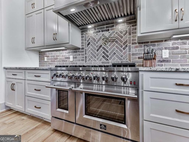 kitchen featuring wall chimney exhaust hood, light stone counters, tasteful backsplash, light hardwood / wood-style floors, and range with two ovens