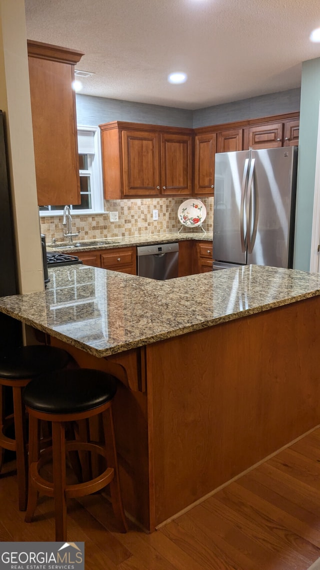 kitchen with light stone counters, kitchen peninsula, stainless steel appliances, hardwood / wood-style floors, and backsplash