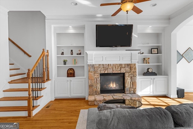 living room featuring built in shelves, a stone fireplace, light wood-type flooring, ornamental molding, and ceiling fan