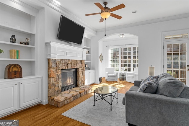 living room with built in shelves, a stone fireplace, light wood-type flooring, ornamental molding, and ceiling fan