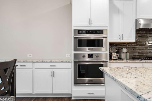 kitchen with white cabinets, backsplash, stainless steel appliances, light stone countertops, and wall chimney range hood