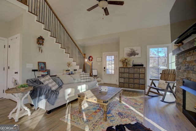 living room with ceiling fan, a towering ceiling, a stone fireplace, and light wood-type flooring