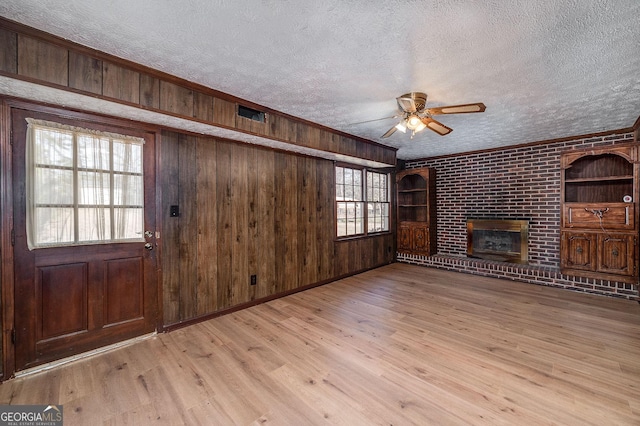 unfurnished living room with light wood finished floors, wooden walls, a fireplace, a textured ceiling, and a ceiling fan