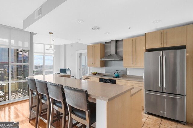 kitchen with pendant lighting, high quality fridge, a center island with sink, wall chimney exhaust hood, and light brown cabinets