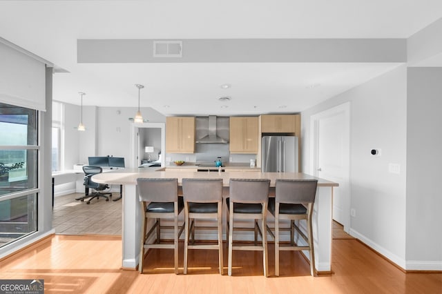 kitchen featuring light brown cabinetry, decorative light fixtures, a kitchen bar, high end fridge, and wall chimney exhaust hood