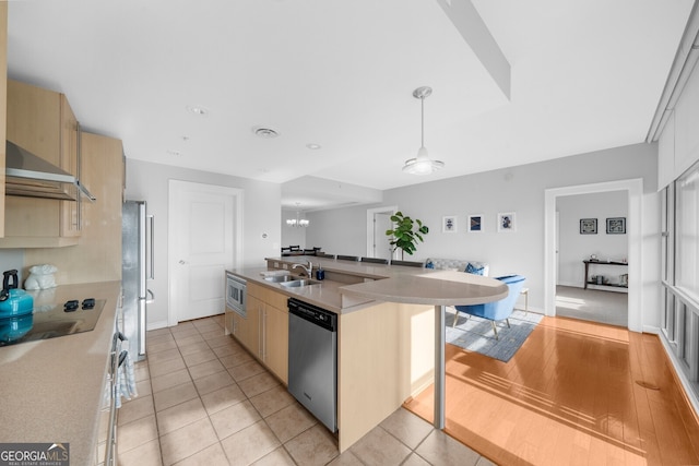 kitchen featuring sink, wall chimney range hood, appliances with stainless steel finishes, hanging light fixtures, and an island with sink