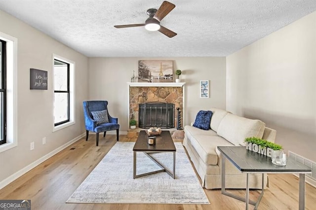 living room with ceiling fan, a stone fireplace, a textured ceiling, and light hardwood / wood-style floors