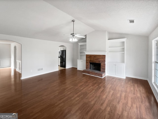 unfurnished living room featuring built in shelves, dark wood-type flooring, lofted ceiling, a brick fireplace, and ceiling fan