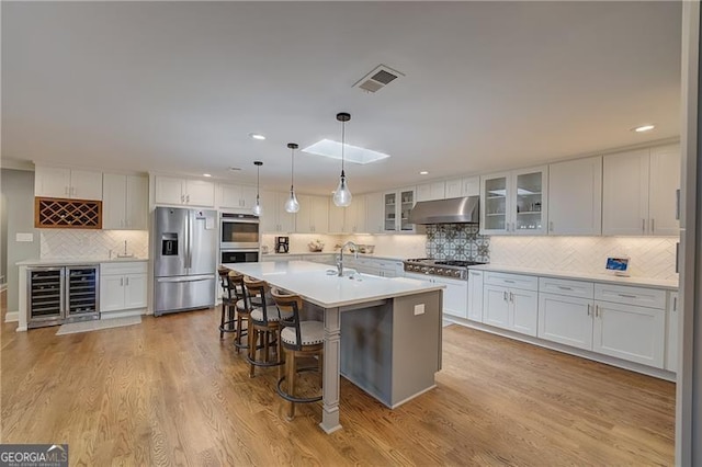 kitchen with appliances with stainless steel finishes, hanging light fixtures, light hardwood / wood-style floors, white cabinets, and a center island with sink