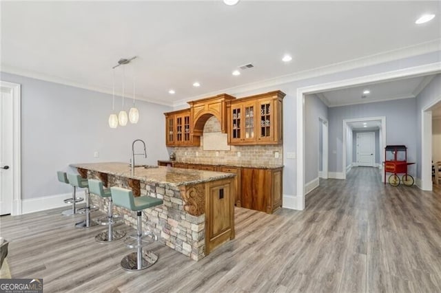 kitchen with pendant lighting, tasteful backsplash, ornamental molding, light stone countertops, and a center island with sink