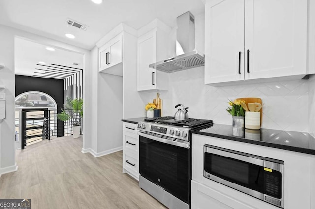 kitchen featuring appliances with stainless steel finishes, white cabinets, backsplash, wall chimney range hood, and light wood-type flooring