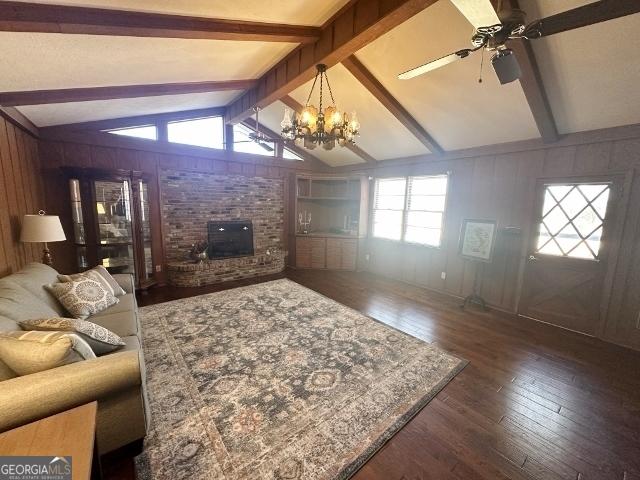 living room with wood walls, lofted ceiling with beams, a brick fireplace, dark hardwood / wood-style floors, and ceiling fan with notable chandelier