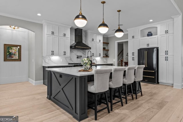 kitchen with high quality fridge, an island with sink, white cabinets, hanging light fixtures, and wall chimney range hood