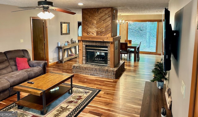 living room with wood-type flooring, a brick fireplace, ceiling fan with notable chandelier, and a textured ceiling