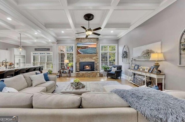 living room featuring ceiling fan, beam ceiling, coffered ceiling, a stone fireplace, and light wood-type flooring