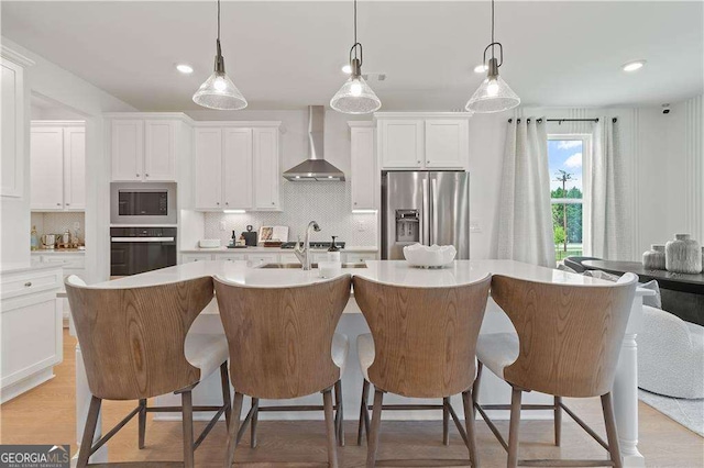 kitchen featuring wall chimney range hood, white cabinets, a center island with sink, stainless steel fridge with ice dispenser, and oven