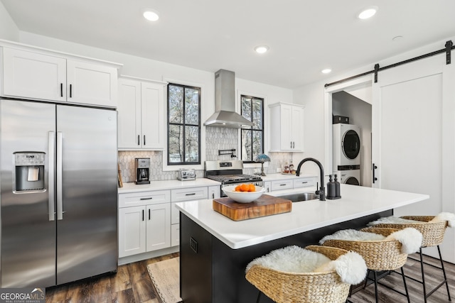 kitchen with appliances with stainless steel finishes, stacked washer / dryer, sink, a kitchen island with sink, and wall chimney range hood