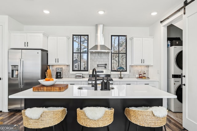 kitchen featuring an island with sink, stacked washing maching and dryer, stainless steel appliances, a barn door, and wall chimney range hood